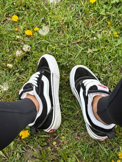 A high-angle, full shot shows a person's feet clad in black leggings and black and white Vans sneakers standing on a grassy field in Teghut, Armenia. The grass is interspersed with yellow dandelions and their white seed heads.