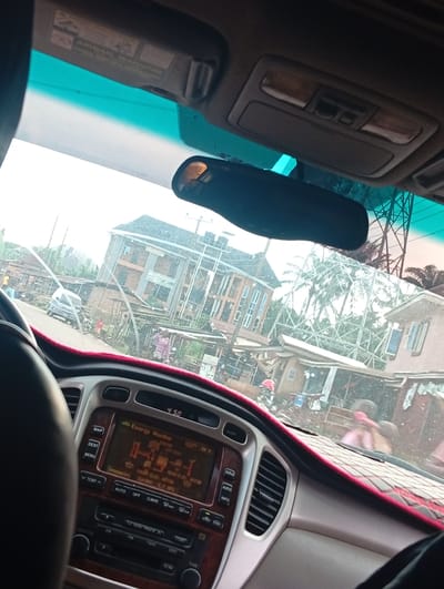 This media file is an image taken from inside a car, looking out through the windshield. The interior of the car is visible, including the dashboard with its radio display showing "Energy Monitor" and the time "4:50". The scene outside the car appears to be a street in Ukpor, Nigeria.
Several buildings and structures are visible, including a multi-story building with an orange facade and a white sign that is partially obscured. There are also smaller structures that suggest a market or commercial area. Palm trees are present, indicating a tropical or subtropical climate.
There are several people visible outside the car, some walking and others standing. A small white car is parked on the street, and there are also motorbikes. The weather appears to be overcast or hazy, and the lighting suggests it might be late afternoon.
The image captures a slice of daily life in what seems to be a bustling urban or semi-urban environment in Nigeria. The focus is on the interplay between the car's interior, the technology it contains (the dashboard display), and the external environment.