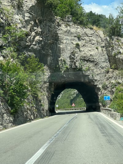 A road winds through a mountainous landscape in the city of Unknown, Montenegro. A tunnel has been bored through a steep, rocky cliff face, allowing the road to pass. A blue road sign is posted near the tunnel entrance, and a warning sign for a sharp curve is visible inside the tunnel. The road itself is gray and appears to have skid marks. Vegetation, including trees and shrubs, clings to the rocky terrain. The sky is visible in the upper left corner.