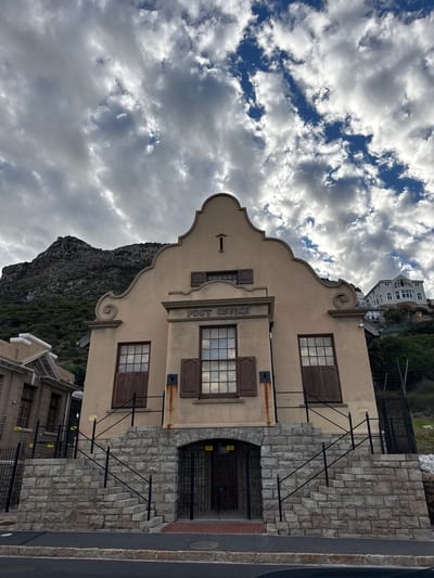 The image shows a historic building that houses the Muizenberg Post Office in South Africa. The building is a two-story structure with a distinctive Cape Dutch architectural style. It features a decorative gable roof, shuttered windows, and a facade adorned with the words "POST OFFICE." The entrance has stone steps and a dark, metal gate. A mountain rises in the background. The sky is filled with clouds. A road is visible on the right side of the building. There are adjacent structures to the sides and to the back of the post office building. The foreground and surrounding areas appear to be relatively empty.