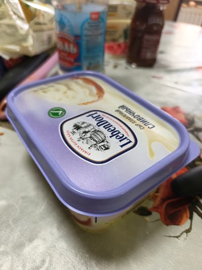 An overhead, slightly angled shot shows a pale purple tub of cream cheese on a patterned tablecloth. The tub has a clear lid, and the cream cheese inside appears to be a pale yellow color with some lighter swirls. On the lid of the tub, a label features the brand name "LiebenDorf" in large, dark letters. Below the brand name, there are smaller words in Russian and German, indicating "СЫР ПЛАВЛЕНЫЙ СЛИВОЧНЫЙ" (melted cream cheese) and "EINFACH GUTER KÄSE" (simply good cheese). There is also an illustration of a carriage on the label. To the left of the tub, partially visible, are some out-of-focus plastic bags and bottles, one of which is a blue bottle with red lettering. To the right, the edge of the tub meets a dark object, likely a piece of cutlery or another kitchen utensil. The tablecloth has a subtle floral pattern. The setting appears to be a domestic kitchen or dining area. There are no people or animals visible in the image. The scene is set in the city of Gavrilkovo, Russia.