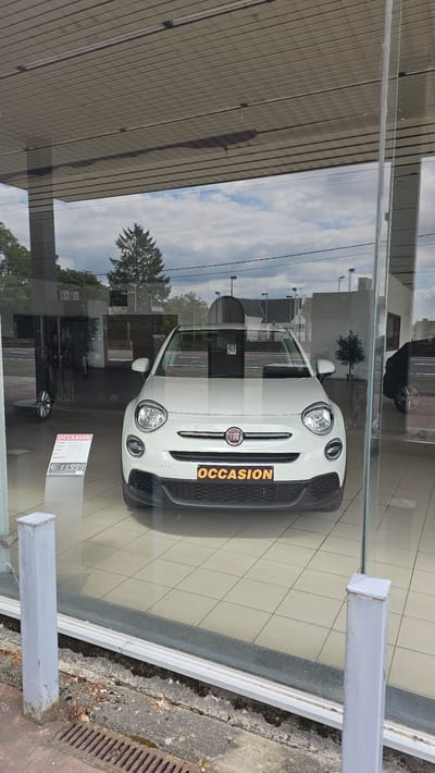 The image shows a white Fiat 500x on display in a car dealership showroom. The car is positioned in the center of the frame and is facing forward. It has a black grille with a Fiat badge in the center and a license plate holder that reads "OCCASION" in orange lettering. To the left of the car, there is a display stand with information about the car, including its price, which is listed as €11,999. The showroom floor is tiled with light-colored squares.
The car dealership appears to be located in a city setting. Through the showroom window, we can see a street with a sidewalk, trees, and buildings in the background. The sky is cloudy. The exterior of the building has a modern design with large glass windows. The area outside the dealership appears to be a road with some vegetation and lampposts. The overall impression is of a well-maintained car dealership showcasing a used car.
Based on the reference provided, the car dealership is located in Boussu-lez-Walcourt, Belgium.