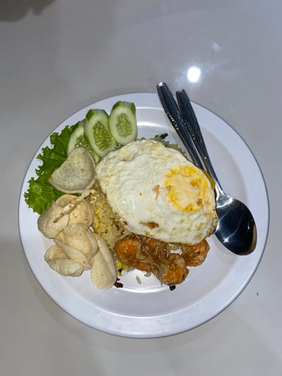 A plate of fried rice with toppings sits on a white table. The fried rice is topped with a fried egg, several cooked shrimp, and some crispy fried shallots. To the side of the rice are a few pieces of lettuce, sliced cucumbers, and some shrimp crackers. Two spoons are placed on the plate, one to the side and one resting on the rice. The setting appears to be an indoor environment, possibly a restaurant or home, given the lighting and white surface. The food is presented as a meal. The image does not contain information about the specific location of Mekar Bakti, Indonesia, nor does it depict any people or actions.