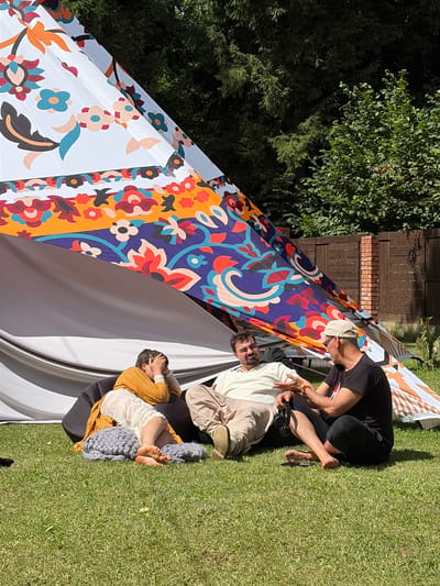 Three individuals are seated on a grassy lawn in front of a large, colorful teepee-like tent. The teepee has a white base with vibrant floral patterns in red, orange, blue, and black. The ground is covered in short, green grass.
On the left, a person is leaning back on a large black beanbag chair, wearing a yellow sweater and a white dress. They have their hands on their head, appearing to be resting or in thought. To their right, a person is sitting with their legs extended, wearing a light-colored long-sleeved shirt and light-colored pants. They are gesturing with their left hand as if speaking. To the right of this person, another individual is seated with their legs crossed, wearing a black t-shirt and black pants. They are looking towards the person in the light-colored shirt and have a baseball cap on.
In the background, there is a wooden fence and trees. The lighting suggests it is daytime with sunlight casting shadows. The overall scene appears to be a casual outdoor gathering. The location is identified as Shebanovo, Russia, implying an outdoor setting within or near the village.