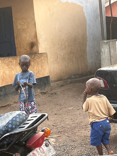 The image shows two young boys outdoors in what appears to be a rural or semi-urban setting. The photo is oriented vertically, with the ground and horizon running from left to right across the image.
On the left side of the frame, a young boy with closely shaven hair is standing, wearing a light blue short-sleeved t-shirt and patterned shorts with blue, white, and red motifs. He is holding two thin, light-colored sticks or pieces of wood in his hands, appearing to be manipulating or playing with them. His gaze is directed downwards at what he is holding.
To the right, another young boy, also with closely shaven hair, is visible. He is wearing a light tan or yellowish short-sleeved t-shirt and blue shorts. He stands with his body slightly angled, looking down towards his left hand or mouth.
In the foreground on the far right, part of a dark-colored motorcycle or scooter is visible. It features a red taillight and an orange turn signal. A metal rack extends from the back of the vehicle, and the seat is covered with a dark material featuring lighter patterned designs, possibly stars or geometric shapes.
The background consists of buildings with plain, light brown or tan walls. There are visible patches and variations in the wall texture, suggesting age or simple construction. A dark-colored door or window shutter is visible on the far left. The ground is unpaved and appears to be dirt or earth, with some areas looking a bit lighter or sandy.
The scene is brightly lit, indicating daytime, likely under direct sunlight. No specific text is clearly legible in the image. The overall atmosphere suggests a casual moment of children playing outdoors in a community.