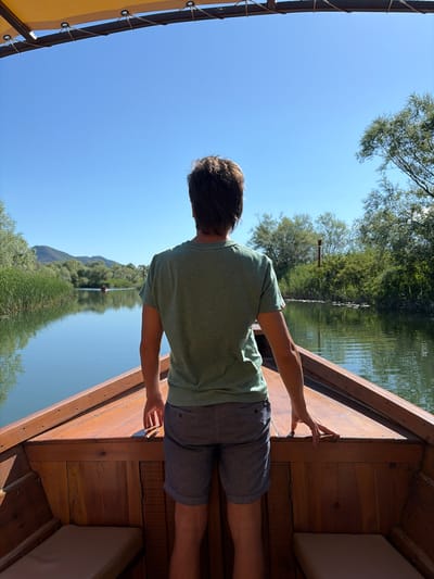 The image depicts a solitary individual, observed from behind, standing on the bow of a wooden boat on a calm river in Braćeni, Montenegro. The person, an adult appearing male, wears an olive green short-sleeved t-shirt and dark grey shorts. Their hands rest lightly on the polished wooden gunwale of the boat, which features internal wooden planking and two light-colored cushioned seats visible on either side. Above, a segment of a yellow fabric canopy with eyelets and securing ropes is visible.
The boat navigates a tranquil waterway, bordered by dense riparian vegetation consisting of tall green reeds and various deciduous trees, with sunlight illuminating the scene. The water surface reflects the clear blue sky and the surrounding greenery. In the far distance, another small boat is visible creating a minimal wake. Low, distant mountains or hills are silhouetted against the clear blue sky on the left horizon. A red-brown marker pole is visible on the right bank amidst the foliage.