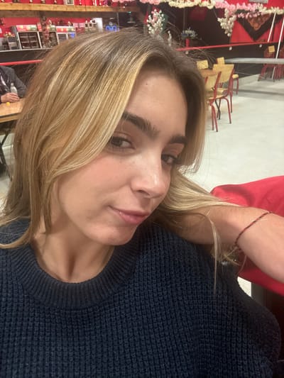 A female subject, approximately 20-30 years old, with light brown and blonde highlighted hair, is positioned centrally, looking directly into the camera with a neutral to slightly smirking expression. She is wearing a dark navy blue knitted sweater with a ribbed crew neck. A thin, braided red and black bracelet is visible on her right wrist, which rests on a red surface, likely a chair back or cushion.
The scene is set indoors in Décines-Charpieu, France, within a venue featuring prominent red walls in the background and mid-ground. Overhead, light-colored garlands, possibly artificial pink and white flowers or draped fabric, are strung across the upper wall area. Additional artificial floral arrangements in white, pink, and blue are visible on the right. Shelves against the red wall display various objects, including what appear to be trophies and other decorative items.
To the left, a portion of another individual's left arm and shoulder, clad in a dark jacket or blazer, is visible, with their hand resting on a light wooden table. Multiple light-colored wooden tables and stackable chairs with red/orange backs and seats are arranged throughout the background on a light-colored, possibly concrete or tiled floor. The lighting is diffused and ambient, suggesting an indoor setting.