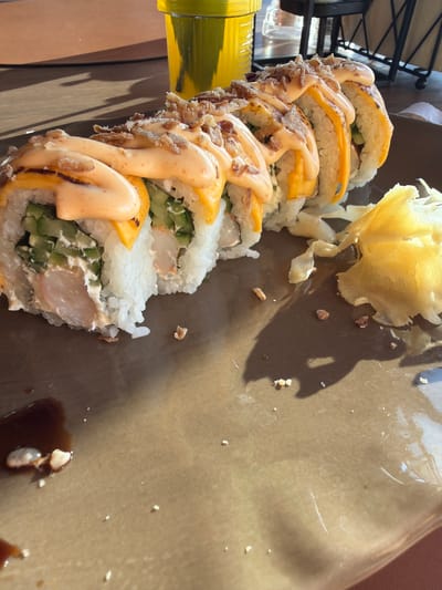 A close-up, high-angle shot captures a plate of sushi with various toppings, including a creamy orange sauce and crispy fried onions, placed on a brown plate. The sushi roll is cut into several pieces and arranged in a diagonal line. To the right of the sushi is a mound of pickled ginger. In the background, a yellow cup filled with a yellow liquid is visible, along with shadows cast on a wooden surface. The scene is set outdoors, likely at a restaurant, given the presence of a table and chairs. There is no indication of the city of Khotianivka, Ukraine, within the image.