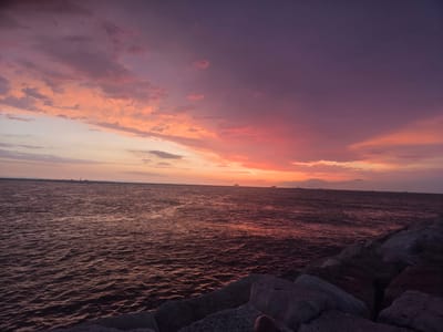 This image captures a vibrant sunset over the Adriatic Sea, viewed from a rocky breakwater in Chioggia, Italy. The sky is a dramatic blend of deep purples and fiery oranges, with scattered clouds tinged pink and yellow. The sea below reflects the colors of the sky, appearing as a rippling expanse of deep red and purple hues. In the distance, several small boats and structures are visible on the horizon. The foreground is dominated by large, rough-hewn rocks, forming a natural barrier against the sea. The overall impression is one of natural beauty and tranquility, with the warm tones of the sunset creating a dramatic and picturesque scene.