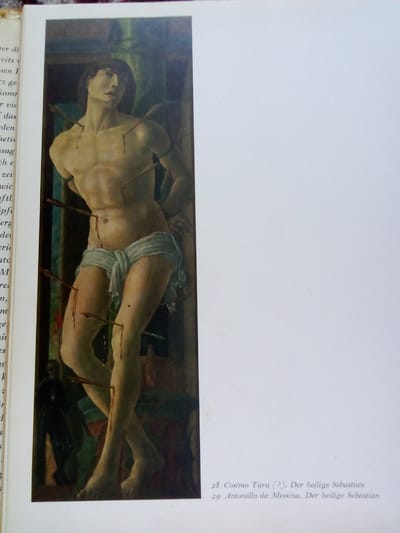 The image is a photograph of a book open to a page featuring a painting of Saint Sebastian. The painting depicts a muscular male figure, likely Saint Sebastian, tied to a post and pierced by arrows. Bloodstains are visible on his body. The figure is partially draped in a white cloth. To the left of the painting is a column of text in German. Below the painting, there is text that reads "28 Cosimo Tura (?). Der heilige Sebastian" and "29 Antonello da Messina. Der heilige Sebastian". The photograph is taken from a slightly elevated angle, showing the white page and a textured, reddish-brown border of the book's cover. There is no discernible connection to Zhmerynka, Ukraine, in the image itself or the accompanying text.