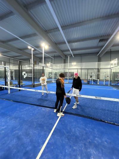 Three men are on a blue paddle tennis court, near the net. One man wears a pink hat and white shorts, holding a black paddle. Another man wears black pants and a black shirt with orange shoulders, also holding a paddle. The third man is wearing a white shirt and gray pants. The court has glass walls and the roof is metal.