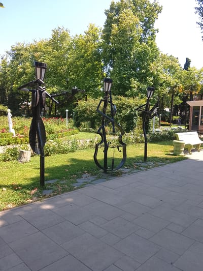 Three abstract metal sculptures resembling musicians are positioned in a park. The sculptures are black and depict figures playing string instruments, with lamp-like structures atop their heads. The sculpture on the left appears to be playing a violin, the center sculpture is designed like a cello, and the sculpture on the right is also playing a string instrument. They stand on a paved pathway in front of a grassy area with rose bushes and trees. A white bench is visible in the background, as well as a small building. The sky is clear and blue, indicating a sunny day. The location is identified as Oryol-Izumrud, Russia.