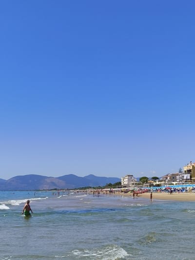 The image depicts a wide, sandy beach in Sperlonga, Italy, under a clear blue sky. The foreground is dominated by the shallow, clear waters of the Tyrrhenian Sea, with gentle waves lapping the shore. A man in green swim trunks stands in the water, facing away from the camera.

In the middle ground, the beach is populated with numerous people, some walking, others sunbathing under colorful umbrellas. Beach chairs and loungers are arranged neatly on the sand. Several small boats are visible near the water's edge.

The background features a strip of buildings, typical of a coastal town, including multi-story structures with balconies and awnings. Green trees punctuate the area between the beach and the buildings. Beyond the town, a range of low, blue-hued mountains forms the horizon. The overall impression is that of a lively and sunny summer day at the seaside.