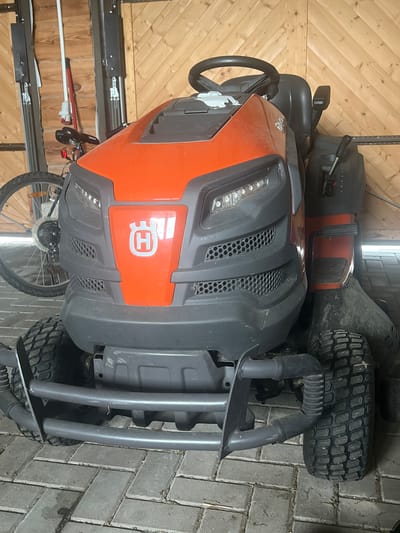 The image displays a garden tractor in what appears to be a garage or shed. The tractor is orange and gray, with a prominent Husqvarna logo on the front. It has large tires and a steering wheel. To the left of the tractor, a bicycle is partially visible. The background consists of wooden walls and a metal door. The floor is paved with brick-like stones. The lighting suggests an indoor environment, possibly illuminated by artificial light. The scene is static and no human activity is depicted. The location in Dubingiai, Lithuania, is not visually evident from the image itself but is provided as context.