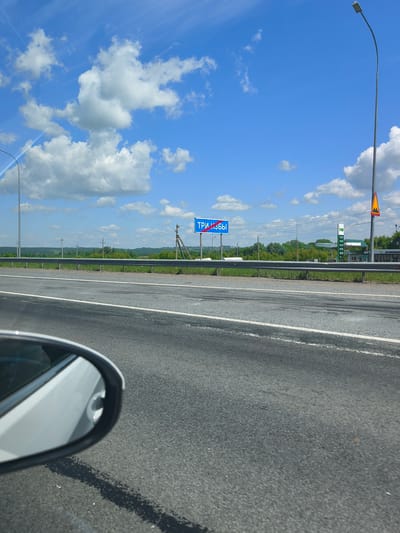 The image shows a view from a car on a sunny day. The road is asphalt with white lane markings. In the distance, there is a blue sign with white Russian text that reads "ТРИ ИЗБЫ" (Tri-Izby) with a red diagonal line through it, indicating the entrance to the settlement. Beyond the road, there is a grassy median strip, a metal guardrail, and a line of trees. Further in the background, rolling hills covered in greenery are visible under a bright blue sky with scattered white clouds. A street lamp is visible on the right side of the image, and a gas station with its price board is also present. The left side of the image shows the side mirror of the car, reflecting the road and the sky. The scene appears to be a rural or semi-rural highway setting in Russia.