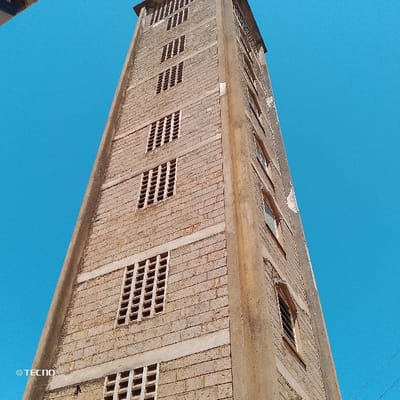 A tall, multi-story building, constructed from light-colored concrete blocks with darker horizontal mortar lines and vertical concrete structural elements, is depicted from a low-angle perspective against a clear, uniform blue sky. This scene is located in Machakos Central ward, Kenya. The left visible facade features at least six distinct levels of rectangular openings, each fitted with a grid of square-shaped concrete blocks or vents. The right visible facade exhibits multiple levels of standard rectangular windows, some with visible frames and reflective glass panes. No individuals, ground features, or surrounding structures are present in the visible environment.