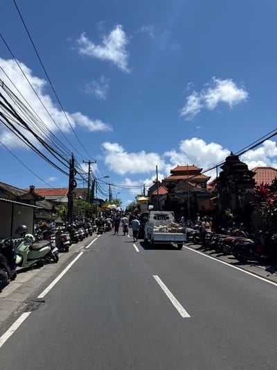 The image depicts a street scene in Kutuh, Indonesia, under a bright blue sky with scattered cumulus clouds. An asphalt road runs through the frame, marked with a white centerline. Parked along one side of the road is a dense line of motorbikes. A white truck is visible on the street, surrounded by several people who appear to be interacting. Traditional Balinese architecture with tiered, terracotta-tiled roofs is present along the roadside. Utility poles with numerous wires crisscross above the street. The overall scene suggests a bustling, everyday environment.