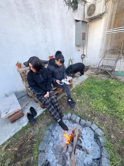 Two individuals are sitting on a wooden pallet next to a white wall. One person is wearing a black jacket and plaid pants, with black socks and a white stripe, resting their feet near a fire pit constructed of stones. Black shoes are placed near the legs. The second person is wearing a black jacket and black pants and is looking at a device in their hands. A black dog sits behind the first person. A fire is burning in the fire pit, which is surrounded by grass. The location is Florida, Argentina.