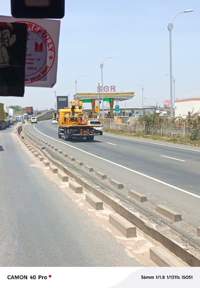 A bright sunny day on a highway shows a yellow Isuzu truck with an attached crane moving away from the camera, nearing the SGR toll station. Concrete barriers line the side of the road. A large sticker with an Isuzu logo, featuring a monkey cartoon and safety reminders, is visible in the foreground. Other cars and trucks are visible in the distance along the highway. The picture was captured with a CAMON 40 Pro camera with the following camera settings: 56mm f/1.8 1/1311s ISO51.