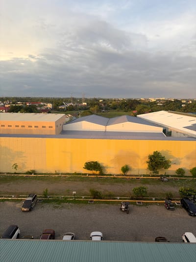 The image shows an aerial view of a section of Dagupan City, Philippines, at what appears to be dusk. In the foreground, a gravel parking lot is visible, with several vehicles parked on it, including a black SUV, a dark red sedan, a silver sedan, and a white sedan. Partially visible at the bottom edge of the frame is the metallic roof of a building.
Behind the parking lot is a long, pale yellow wall, which extends across the width of the image. This wall is bordered by a strip of grass and then a row of trees and shrubs. Further back, several industrial-style buildings with light-colored, sloped roofs are visible. One building has a light beige facade with regularly spaced, small rectangular openings. Beyond these structures, the landscape opens up to a wider view of the city, characterized by a mix of smaller buildings, residential houses with red roofs, and abundant greenery. In the distance, two telecommunications towers are prominent against the horizon.
The sky above is filled with dramatic clouds, with a lighter, potentially sunlit area visible towards the left, suggesting the sun may be setting or rising. The overall atmosphere is muted, with the yellow wall catching the light and casting shadows. No people are directly visible in the scene.