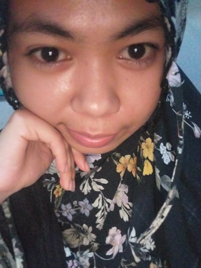 The media file is a close-up, slightly angled selfie of a young person wearing a black hijab with a floral pattern. The background is a blurry, light blue surface, possibly a bedsheet. The person is resting their head on their hand, with their chin and cheek supported by their fingers. Their eyes are open and looking towards the camera, and they have a gentle, relaxed expression. The hijab is dark with a repeating pattern of various flowers in shades of white, yellow, pink, and purple. The lighting suggests an indoor setting, possibly during the day given the brightness. No specific time of day or weather cues are discernible. There is no visible text in the image. The overall impression is one of quiet introspection or a casual, at-home moment. The location context of Watampone, Indonesia, is external information and not directly observable from the image itself.