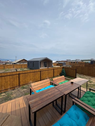 A wide-angle, high-angle outdoor shot captures a wooden deck with a dining table and benches in the foreground, looking towards a residential area in Khuzhir, Russia. The deck is furnished with two wooden benches, one with blue cushions and the other with green cushions, and a large wooden dining table with a light-colored tabletop and dark metal legs. A small ashtray sits on the table. Beyond the deck, a brown wooden fence encloses a grassy yard. In the background, several modern, modular houses are visible, some with dark gray siding and metal roofs. One house features a light brown exterior and a dark roof. A small, artificial Christmas tree stands to the left of the frame. Solar panels are mounted on the roofs of some of the buildings. The sky is overcast with light gray clouds. In the distance, faint mountains can be seen. The scene depicts an outdoor recreational area in a suburban setting.