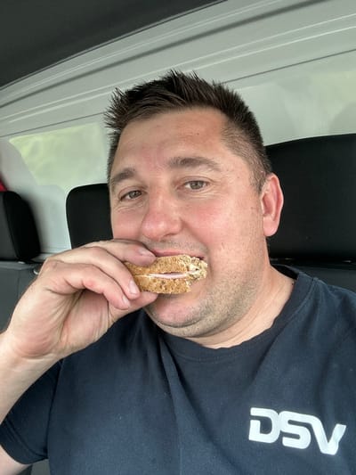 The image shows a close-up of a man holding a sandwich to his mouth, looking directly at the camera. He appears to be in the process of taking a bite or about to eat the sandwich, which is made with brown bread and has a visible pink filling, possibly ham.
The man has short, dark hair, light-colored eyes, and light stubble on his face. He is wearing a dark, short-sleeved T-shirt. The T-shirt has the white, stylized letters "DSV" printed prominently on the chest, visible in reverse due to the image's orientation.
The background behind the man is largely out of focus, consisting of a dark, textured surface, likely a seat or chair, and a light-colored, vertically paneled wall or surface. The setting appears to be indoors. The lighting is even, suggesting a daytime scene. The man's expression is neutral and direct towards the camera. There are no explicit visual cues within the image to confirm the provided location context of Wädenswil, Switzerland.