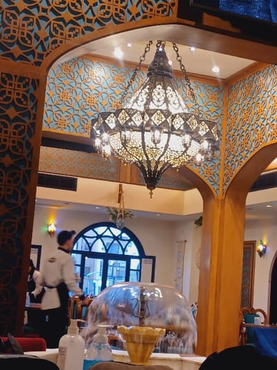 The image depicts the interior of a restaurant or large dining hall in Dummar Municipality, Syria, characterized by traditional Middle Eastern or Islamic architectural and decorative elements. A prominent, ornate metal chandelier with multiple illuminated bulbs hangs centrally from a decorative arched ceiling. The surrounding walls and archways feature carved wooden frames and light blue panels with intricate gold geometric patterns. In the mid-ground, a circular water fountain or feature with a transparent, dome-like cover is visible, with water flowing over its surface, and a smaller golden pedestal in front.

To the left of the fountain, a male staff member in a light-colored long-sleeved shirt, a dark apron, and a dark cap walks away from the camera, heading towards a large arched window or doorway that reveals an exterior building structure. A partially obscured figure is visible behind him. In the immediate foreground, on a table, two pump bottles—one white, one blue—likely containing hand sanitizer, are present. The overall lighting is warm, emanating from the central chandelier, recessed ceiling lights, and accent lighting on the walls, illuminating a space designed with elaborate details and a welcoming ambiance.