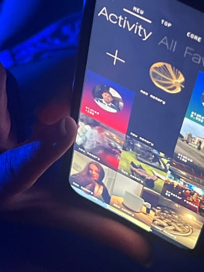 The image shows a smartphone screen displaying an app interface with the word "Activity" prominently featured. The app seems to be centered around content creation, as indicated by the terms "creator redeen," "staked," and "new nenory," which may refer to the monetization or staking of content. The screen also displays thumbnails of various images and videos, likely representing user-generated content within the app. A person is holding the phone, and the background suggests a dark or dimly lit environment, possibly indoors. The lighting seems to be blue. The image is rotated. There is no evidence to suggest that this photo was taken at Heenatigala Junction, Sri Lanka.