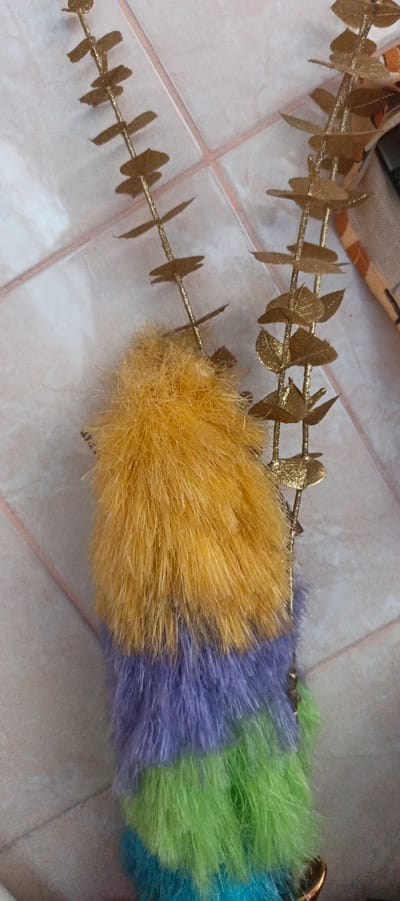 The image displays a close-up view of a decorative item, likely a garland or a string of ornaments. Two golden, glittery stems with leaf-like embellishments are prominently featured. Hanging from these stems is a fluffy, multi-colored tassel. The tassel transitions from a vibrant yellow at the top, followed by sections of purple and then green. The background appears to be a tiled surface, possibly a floor or wall, with a light-colored, possibly cream or beige, tone and faint pink grout lines. The lighting suggests an indoor setting. There is no information or visual evidence present to indicate the location in Tikhvin, Russia, or any other specific geographical context. The image focuses solely on the details of the decorative object and its immediate surroundings.