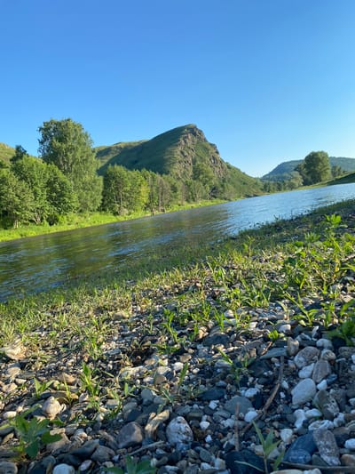 A bright, sunny day in Bol'shearslangulovo, Russia, showcases a tranquil river flowing through a lush, green landscape. The foreground is a stony bank covered with sparse green grass. The river, reflecting the clear blue sky, meanders towards the horizon, bordered by a dense line of trees on the left. Beyond the trees, rolling hills rise, with one prominent peak catching the sunlight. The overall scene is one of natural beauty and serenity.