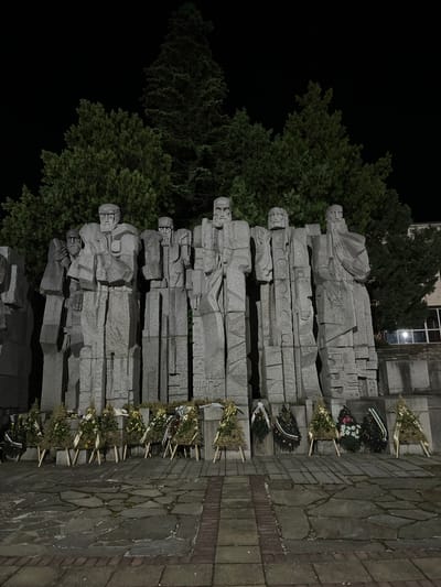 This nighttime, low-angle photograph captures a prominent stone monument in Veliki Preslav, Bulgaria, featuring a row of at least seven stylized, blocky male figures. The monument, rendered in grey stone with an angular, Brutalist aesthetic, depicts the figures standing upright with solemn expressions and indistinct, flowing attire, illuminated by artificial light.
In the immediate foreground, arranged along the base of the monument, are numerous commemorative wreaths. These include both triangular wreaths, primarily composed of evergreen branches and decorated with ribbons (some golden, others dark or featuring national flag colors), and circular wreaths. Each wreath rests on a small, light-colored wooden stand. The ground beneath is paved with irregular dark stone slabs and bordered by brickwork, showing signs of dampness.
Behind the monument, the background is dominated by a dense array of tall evergreen trees, appearing dark against the black night sky. On the far right, partially visible, is a building with illuminated windows, suggesting an urban or semi-urban context. The scene conveys a sense of solemn commemoration.