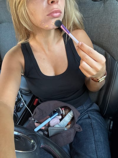 A female individual, located in Cidade da Maia, Portugal, is depicted inside a vehicle, applying makeup. She is seated, wearing a black ribbed tank top and dark denim pants. Her blonde hair falls over her shoulders. Visible on her chin and lower cheek are several distinct spots of beige-toned liquid foundation. She holds a makeup brush, featuring a black head, purple ferrule, and white handle, in her right hand, positioned near her chin as if blending the foundation. A brown leather-strapped wristwatch with a gold-colored rectangular face is worn on her left wrist. On her lap, between her legs and the steering wheel, rests an open dark brown textured makeup bag filled with various cosmetic items, including multiple brushes (one with a light blue handle), compacts, and tubes. The car's interior features dark grey fabric seats with visible stitching. To the left, a portion of a dark steering wheel with textured grip and red stitching is visible. Further left, elements of the car's central console or dashboard are discernible, including control buttons and a white charging cable connected to a port.