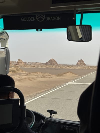 The image depicts a view from inside a vehicle, likely a bus, looking out onto an arid desert landscape. The perspective is from the driver's or front passenger's seat, with parts of the steering wheel, dashboard, and a rearview mirror visible on the right. A paved road with white lane markings runs parallel to the vehicle, alongside a vast expanse of sandy desert.

In the distance, unique natural rock formations, appearing as weathered mesas or yardangs, stretch across the horizon under a hazy, overcast sky. The overall scene suggests travel through a stark, expansive desert region.

Notable details include a green-tinted section of the windshield and a black sun visor in the upper left. The sun visor clearly displays the text "GOLDEN DRAGON" in silver lettering, accompanied by a circular logo, indicating the brand of the vehicle. The head and shoulder of a person, presumably the driver or a passenger, are visible in the lower right corner of the frame. The time of day appears to be daytime, possibly under diffused sunlight due to the hazy conditions. The setting is consistent with the desert environment of Dunhuang City, China.
