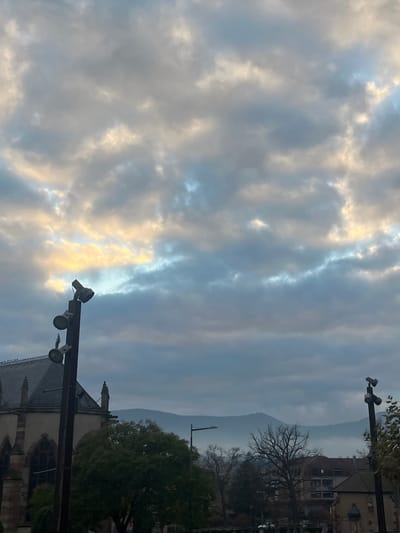 The image captures an outdoor urban landscape in Obernai, France, under an overcast sky. The sky is largely covered by dense, textured clouds, exhibiting varying shades of grey, blue, and white, with prominent areas of golden-yellow light visible through gaps on the left and upper-left, suggesting indirect illumination.

In the left foreground, a tall, dark metal light pole equipped with three downward-facing spotlights stands prominently. Behind and to the left of this pole, the upper portion of a large stone building with a steeply pitched dark roof is visible. This structure is identified as the Church of Saints Peter and Paul due to its Gothic architectural style, characterized by pointed arch windows and buttress-like features.

To the right of the church, a green deciduous tree with dense foliage occupies the mid-ground. Further into the mid-ground, a modern street lamp with a single horizontal light fixture is visible, positioned in front of a distant mountain range. Another dark metal pole, fitted with two downward-facing spotlights, is partially visible on the far right of the frame. Scattered bare trees and several multi-story buildings with dark roofs and light-colored facades are present in the mid-ground to the right. The background features a hazy, muted silhouette of mountains, consistent with the Vosges mountain range surrounding Obernai. No individuals, overt actions, or interactions are discernible within the scene.