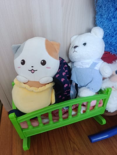 The image shows a green plastic toy crib with two stuffed animals inside. On the left is a calico cat plush toy with a yellow blanket. On the right is a white teddy bear plush toy wearing blue overalls. The crib is placed on a wooden floor next to a light-colored wall. In the background, parts of other plush toys, including a blue one and a white one with blue eyes, are visible. There are no people in the image. The scene appears to be indoors. There is no indication of the city of Soroca, Moldova.