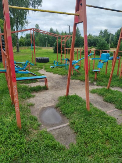 The image shows an outdoor fitness equipment area in Drezna, Russia. The equipment is made of red and blue metal frames, with some yellow bars. There are monkey bars, a bench press, a seesaw, and other exercise machines. The area is surrounded by grass and trees. The ground is a mixture of dirt paths and concrete. There is a puddle of water on one of the paths. The sky is cloudy. No people are visible in the image.