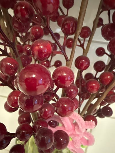 A close-up, high-angle shot shows a cluster of dark red, artificial berries on thin, brown stems. The berries are round and have a glossy finish, reflecting light. Some are in sharp focus, while others are blurred. In the lower right, a portion of a pink, fuzzy object, possibly a craft flower, is visible. The background is a pale, out-of-focus white. There are no people or animals in the image. The scene is devoid of any specific environmental context that would indicate a location in Ridabu, Norway, or any other city.