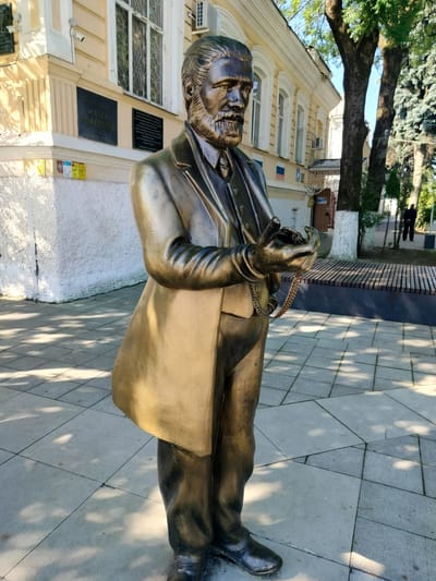 A bronze statue of a bearded man in a suit and overcoat stands on a paved walkway in front of a light-colored building with arched windows. The statue is positioned in the foreground, viewed from the knees up, with his hands held together in front of him. He appears to be holding a pocket watch in his right hand, attached to a chain. The building behind him has several plaques on its facade, one of which reads "МУЗЕЙ" (MUSEUM) in Russian. Trees with light-colored trunks and dark green foliage are visible to the right of the building, casting dappled shadows on the walkway. In the background, a person in dark clothing is walking away from the camera on another paved path. The setting appears to be an outdoor public space, likely in the city of Labinsk, Russia, given the context.