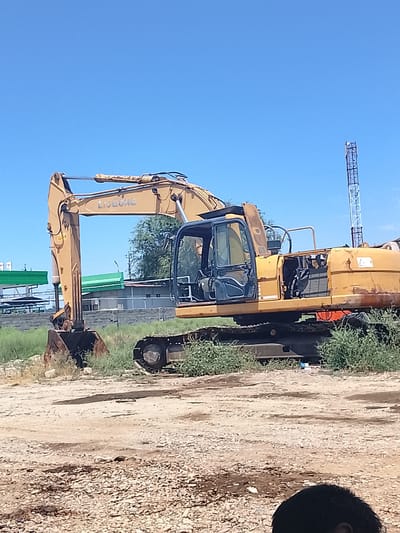 A large yellow excavator sits parked on a dirt lot. The excavator is the main subject, with its boom extended and bucket lowered. It appears to be an older model, with some rust visible on its body. The cab is open, revealing the interior. To the left of the excavator, there are some green structures that resemble gas station canopies. In the background, there is a tall metal tower, possibly a cell tower, and some trees. The sky is clear and blue, suggesting a sunny day. The ground is covered in dirt, gravel, and some sparse vegetation. A portion of a person's head is visible in the foreground, suggesting this is a photograph taken by someone present at the scene. The overall impression is that of an industrial or construction site, possibly abandoned or in disuse, under a bright, clear sky. The excavator has the brand name "LIUGONG" visible on its boom. The setting appears to be an outdoor location in Sabir Rakhimov, Uzbekistan.