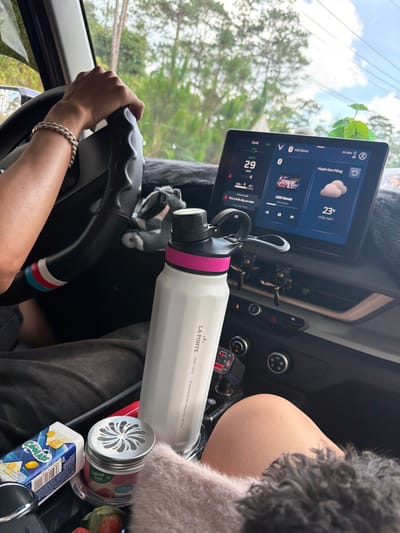 The image captures an inside view of a car, likely in Măng Đen, Vietnam, focusing on the dashboard and parts of the front cabin. A person's hands are visible on the steering wheel, suggesting they are driving. Another person's leg and a furry item (possibly a pet or blanket) are seen on the passenger side.

The central infotainment screen displays several pieces of information: the location "Huyện Kon Plông" (Kon Plông District, where Măng Đen is situated), a weather icon indicating clouds and a temperature of "23° mây cum" (23°C cloudy). Music, "H2O Remix," is shown playing, and a speedometer indicates "29 km/h". A safety message, "Chú ý thức dậy an toàn" (Pay attention, wake up safely), is also visible.

Various objects are arranged around the car's interior. A white "LA FONTE 709ML / 24OZ STAINLESS STEEL INSULATED BOTTLE" with a pink and black cap is prominently placed. Nearby, a small stuffed animal, possibly a badger or raccoon, sits on the dashboard. A USB charger with a red digital display showing "0.0" is plugged in. Snacks are visible on the passenger side, including a carton of "COOLAIR" juice and a container labeled "SAMEL Natural Fresh." Several strawberries are also present. The driver wears a silver-colored bracelet on their right wrist. The setting suggests a daytime journey under partly cloudy skies.