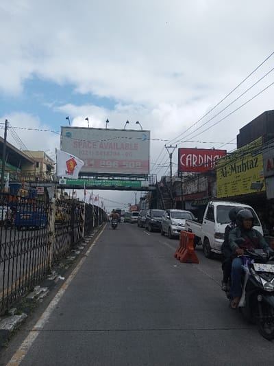 This image depicts a bustling urban street scene in the city of Thotagamuwa, Sri Lanka, under a partly cloudy sky. The perspective is from the roadside, looking down a two-lane asphalt road.
In the immediate foreground on the right, a black motorbike with two male occupants is visible, moving towards the viewer. The driver wears a dark jacket and a black helmet, while the passenger behind him is partially obscured. An orange plastic road barrier sits on the asphalt next to the motorbike. On the left side of the road, a dark metal fence with vertical bars lines the street, separating it from an adjacent area or elevated structure. Several small white flags, each displaying a red political party logo resembling a stylized bull's head, are affixed to this fence.
The road extends into the midground, where a line of vehicles is either parked or slowly moving on the right side. This queue includes a white compact pickup truck, a silver minivan, and several dark-colored cars. Further down the road, a single individual on a motorbike is seen traveling away from the viewer.
Above the street, a large rectangular billboard dominates the upper midground, displaying the text "SPACE AVAILABLE" and two sets of contact numbers: "(021) 5418704 - 06" and "011 896 508". The billboard is illuminated by spotlights mounted on its top edge. Directly below this billboard, a green banner spans the width of the street, carrying text partially visible as "BAYARILAH PAJAK BUMI & BANGUNAN KOP ANDA SEBELUM JATUH TEMPO". To the right of the billboard, a prominent red sign with white lettering displays the word "CARDINAL." Further right, a yellow sign with "AL-MUHTAZ" is visible above a storefront. An elevated pedestrian bridge structure with visible stairs ascending from the right side of the street is also present.
Buildings, likely commercial establishments, line both sides of the street, extending into the background. Overhead power lines crisscross the upper portion of the frame. The sky is predominantly light gray with patches of white and blue clouds. The road surface is black asphalt, bordered on the left by a white solid line.