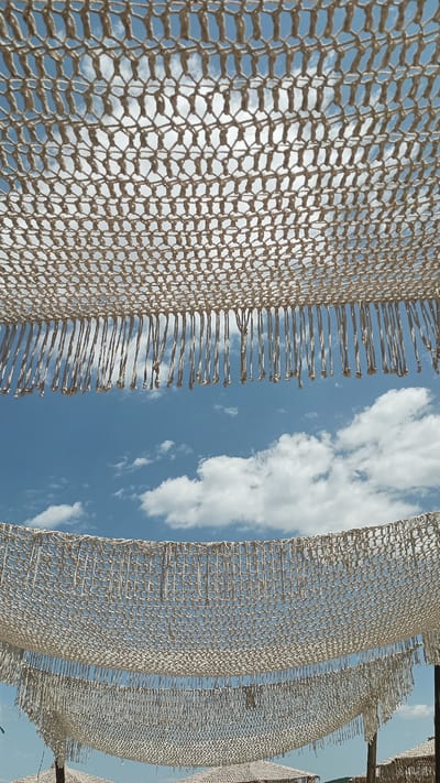 The image depicts two macrame canopies with fringe hanging from them, set against a bright blue sky with scattered white clouds. The canopies are made of thick, off-white rope woven into a diamond pattern. The top canopy is closer to the viewer and appears larger, with a dense fringe of knotted cords. Below it, a second, similarly constructed canopy is visible, also with a fringe of hanging ropes. The bottom of the image shows hints of what might be beach umbrellas or similar structures beneath the second canopy. The lighting suggests it is daytime, likely sunny, given the bright sky and the clarity of the scene. The overall impression is one of a relaxing, possibly seaside or outdoor resort setting in Nea Peramos, Greece. There is no visible text in the image.