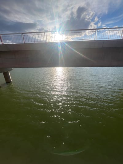 The image depicts a body of water, likely a river, under a bridge in Ait Mazigh, Morocco. The left side shows a section of the bridge structure, including a railing and concrete support. Sunlight streams through the railing, creating a bright glare. The sky is visible through the railing, with scattered clouds. The water surface reflects sunlight, creating a shimmering effect. A portion of a bridge support extends into the water. The water appears green in color. There are no people visible in the image.
