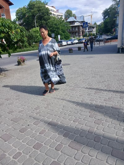 A woman in a gray and white tie-dye dress walks on a cobblestone sidewalk in Truskavets, Ukraine. She is carrying a bag with a black and white pattern over her shoulder and is wearing dark sandals. The sidewalk is bordered by a road with parked cars and a tree-lined area. In the background, there are buildings, including a hotel, and a construction crane. The sky is partly cloudy.