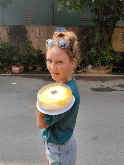 A woman is standing outdoors, holding a cake. She is fair-skinned, with blonde hair pulled up in a messy bun, and is wearing sunglasses on top of her head. She is wearing a dark green t-shirt and light blue jeans. She has her tongue sticking out slightly. The cake she is holding is round, yellow, and in a clear plastic container with a white base. The cake has a blue label with white text on it. She is standing on a gray asphalt street. Behind her is a low building with a faded green wall, with a few windows. In front of the building are various potted plants and trees. Based on prior knowledge, the city of Thành phố Hà Giang, Vietnam, is likely in the background.