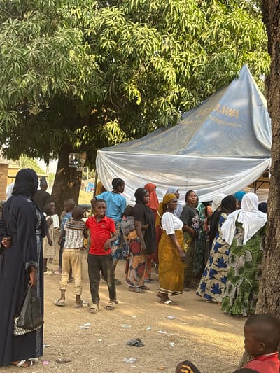 A group of people of varying ages stand outdoors near a large tree and a plastic-covered structure. Most are facing towards the left of the frame. The individuals in the group are dressed in a variety of colorful clothing, including patterned dresses, solid-colored shirts, and traditional garments such as a hijab. Several children are also present. The ground is strewn with debris.