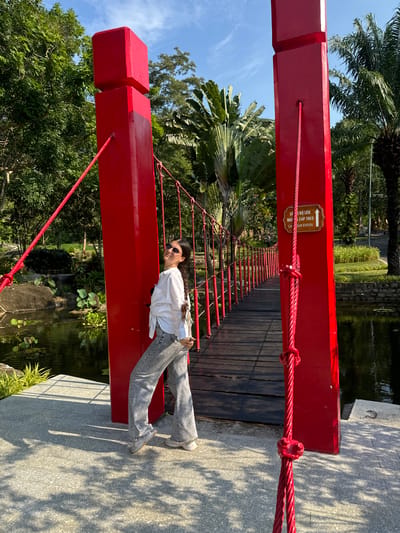 A young woman with long dark hair and sunglasses is posing on a red suspension bridge. She is wearing a white button-down shirt and grey flared jeans. The bridge is made of red painted beams and wooden planks, with red cables as railings. The bridge spans over a body of water surrounded by lush greenery, including palm trees and other tropical plants. The scene suggests a sunny day, possibly in the late morning or early afternoon, given the shadows cast by the trees on the bridge's walkway. The woman has a relaxed and confident demeanor, looking towards the camera with a slight smile. A sign on the bridge reads "LÊN CÁP TREO" and "CAP STATION" in Vietnamese and English, indicating it's a cable car station. The overall impression is a pleasant outdoor excursion in a tropical setting.