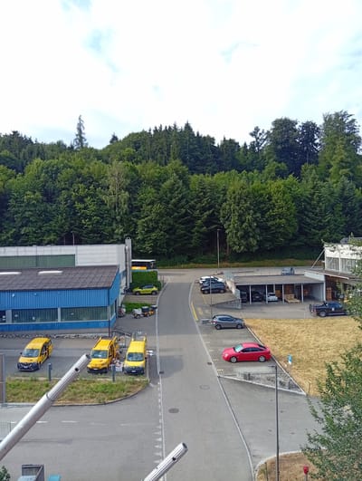 The aerial perspective captures a mixed-use industrial and natural landscape in Zofingen, Switzerland. In the foreground, a gray asphalt road curves, bordered by grass and gravel. Three yellow utility vans, each with a white logo on the door, are parked in a row on the grass to the left. A blue industrial building with a dark gray roof runs along the left side of the image, featuring large windows. Behind it, a light gray building with a modern facade extends further.
The road continues to the right, where a variety of vehicles are present. A yellow hatchback is parked on the grass, and further along, a red sedan is parked in a designated spot. Adjacent to this, a covered parking structure houses several cars, including a white hatchback and a few dark sedans. A black pickup truck with a bike rack is parked to the right of the covered area, near a building with a white facade.
The background is dominated by a dense forest of mature green trees, creating a natural backdrop against a pale sky with scattered clouds. A yellow and orange bus is visible on a road disappearing into the trees in the upper right portion of the image. The overall scene suggests a functional industrial zone situated at the edge of a natural wooded area.