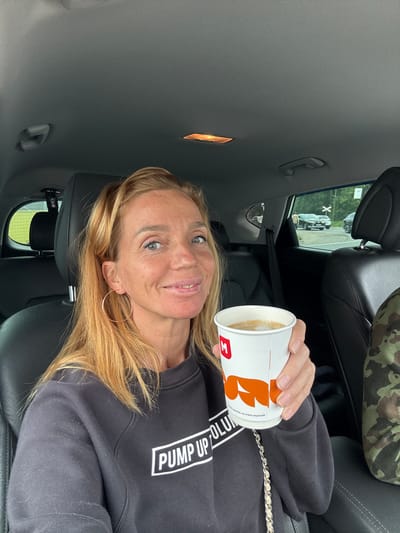 A woman with blonde hair is in the driver's seat of a car, holding a coffee cup in her right hand. She is smiling and looking at the camera. She is wearing a black sweatshirt with white text that reads "PUMP UP OLUN". The car interior is visible, with black leather seats and the headliner above. Outside the car window, a rural scene with trees and a dirt road can be seen. The coffee cup has a logo with an 'M' and orange graphic. The image appears to be taken during the day. The location is the city of Kuivozi, Russia.